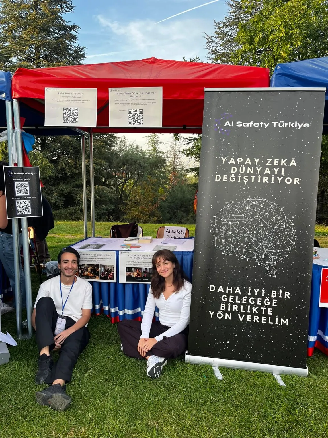 Bilkent PhilFest’26’daki AI Safety Türkiye standının önünde çimlere oturmuş iki kişi; yanlarında “Yapay zekâ dünyayı değiştiriyor, daha iyi bir geleceğe birlikte yön verelim” yazılı afiş
